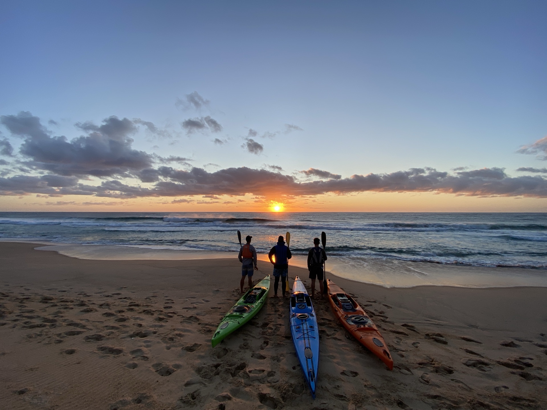 Three kayaks lined up on the sand at sunrise as the crew looks out over the surf