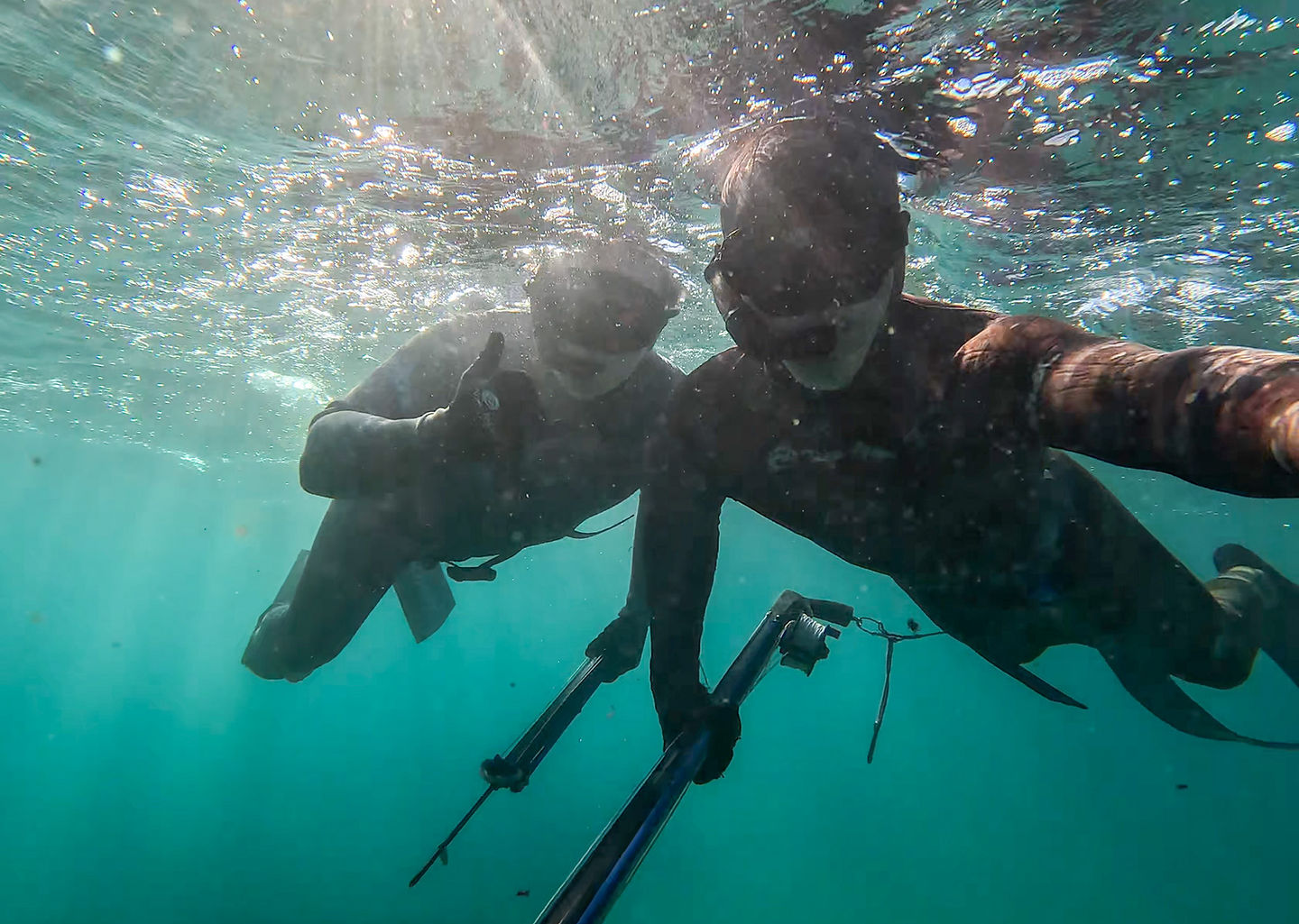 Two spearfishermen at the surface in wetsuits and masks, one giving a thumbs-up, spearguns in hand